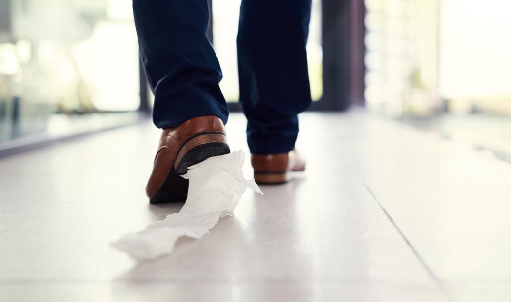 A man in a navy suit and brown brogues strides down a bright office corridor with a strip of toilet paper stuck to his heel.